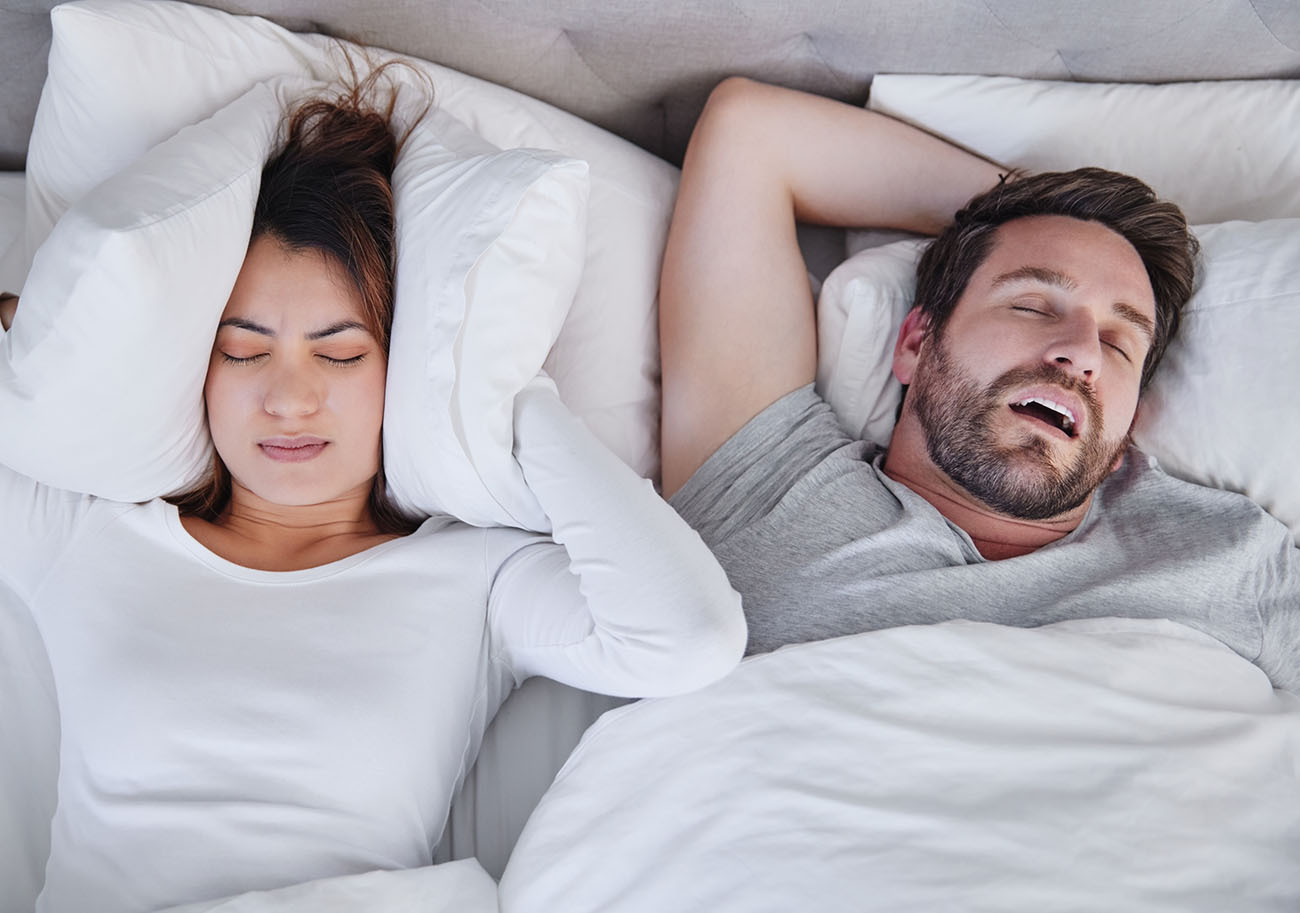 Snoring and sleep apnea can be a symptom of TMJ disorder as seen her with a man snoring and the woman covering her ears with her pillow in Bel Air, Los Angeles.